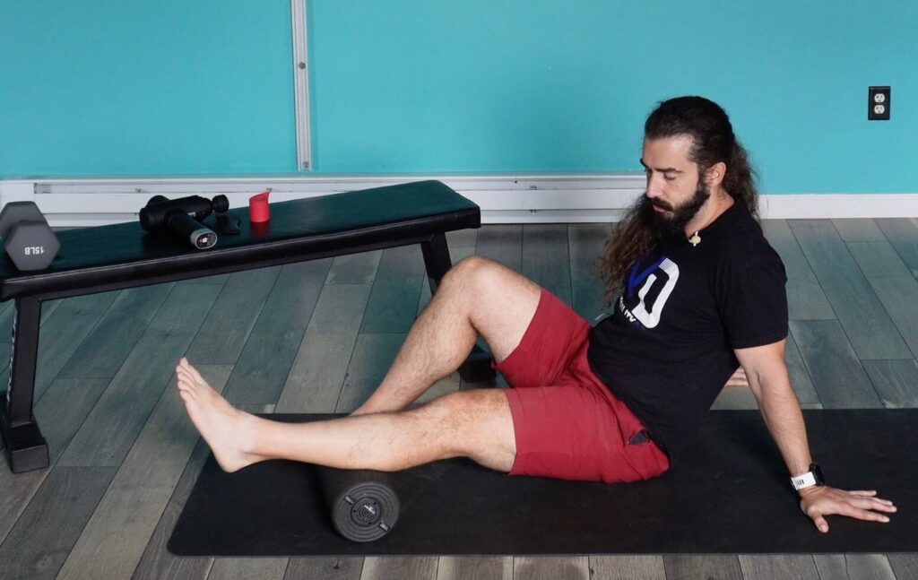 Dr. John seated on the ground supporting himself with his hands with a foam roller under the left achilles tendon